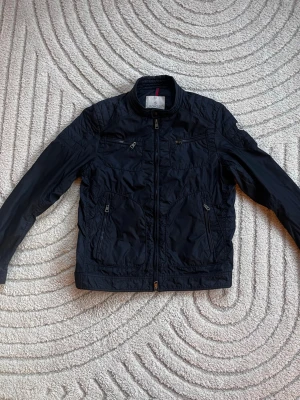 Svart jacka  från Moncler - Snygg svart vindjacka från Moncler med hög krage och dragkedja framtill. Jackan har flera fickor med dragkedja och ett broderat Moncler-märke på ärmen. Tillverkad i lätt, glansigt material som ger en sportig look. Perfekt för blåsiga dagar. Jackan är i stolek 4 