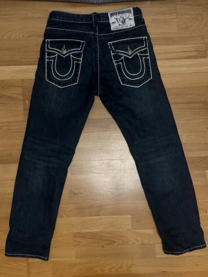 True Religion mörkblå jeans - Mörkblå jeans från True Religion med tydliga vita kontrastsömmar och stora bakfickor med ikonisk brodyr. Klassisk femficksmodell och patch med logga bak i midjan. Snygg och avslappnad passform, perfekt till streetwear-stil.