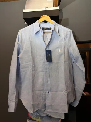 Ljusblå skjorta från Polo Ralph Lauren - Klassisk ljusblå skjorta från Polo Ralph Lauren med lång ärm och vit broderad logga på bröstet. Tillverkad i bomull och har knappar framtill. Perfekt för en clean och stilren look.