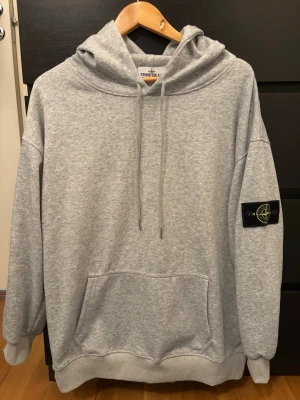 Grå hoodie från Stone Island - Snygg grå hoodie från Stone Island med klassisk patch på vänster ärm. Hoodien har en huvficka Tillverkad i mjuk bomullsblandning som är skön att ha på sig. Perfekt för en avslappnad streetwear-look.