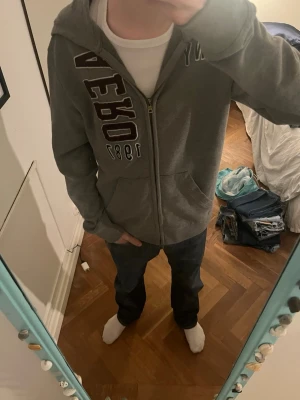 Grå zip-hoodie från Aeropostale - Snygg grå hoodie med dragkedja från Aeropostale. Stor tryckt logga i mörkrött och vitt på framsidan samt årtalet 1987. Klassisk huva och två fickor framtill. Perfekt för en avslappnad och cool stil. Med en fett fläck som jag är osäker på om den går bort