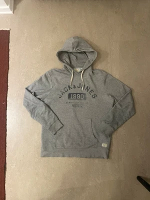 Grå hoodie från Jack & Jones - Grå hoodie från Jack & Jones med tryck framtill och vita dragsnören i huvan. Klassisk känguruficka och ribbade muddar vid ärmslut och nederkant. Perfekt för en avslappnad stil.