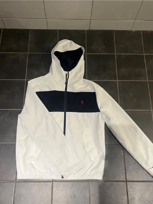 Vit vindjacka från Polo Ralph Lauren - Vit vindjacka med svart panel över bröstet och röd Polo-logga. Jackan har huva, dragkedja framtill och två fickor. Perfekt för blåsiga dagar och har en sportig, stilren look. Pris kan diskuteras!!