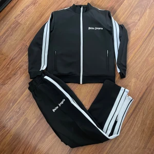 Palm Angels svarta mjukisbyxor med ränder - Svarta mjukisbyxor från Palm Angels med vita sidoränder och logga på benet. Byxorna har elastisk midja och dragkedja vid benslut. Tillverkade i polyester för en sportig vibe. Perfekta för en streetwear-look.