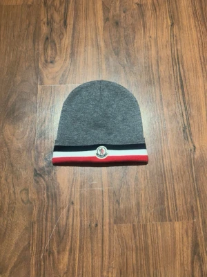 Grå Moncler mössa med ränder - Snygg grå mössa från Moncler med klassiska ränder i svart, vitt och rött längs kanten. Mössan har Moncler-loggan framtill och är tillverkad i mjukt stickat material. Perfekt accessoar för att lyfta din stil under kyliga dagar.