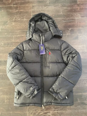 Svart pufferjacka från Polo Ralph Lauren - Snygg svart pufferjacka från Polo Ralph Lauren med avtagbar huva och dragkedja framtill. Jackan har två sidofickor med dragkedja och ikonisk röd logga på bröstet. Perfekt för kalla vinterdagar och riktigt skön att bära.