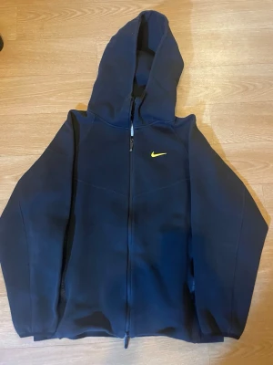 Svart Nike Nocta tracksuit med gul Nike logga! - Svart Nike tracksuit från Nike, dragkedja och huva. Liten gul Nike-logga på bröstet och diskreta gula detaljer på ryggen. Tillverkad i mjukt material och har långa ärmar. Perfekt för en sportig och avslappnad stil. 