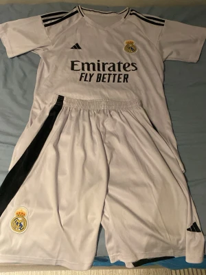 Real Madrid matchställ Adidas L - Säljer ett vitt Real Madrid matchställ från Adidas med svarta detaljer. Tröjan har klubbmärke och sponsortryck framtill samt klassiska Adidas-ränder på axlarna. Shortsen har klubbmärke och svart sidopanel. Perfekt för fotbollsträning eller match.