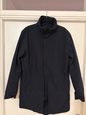 Scandinavian Edition - Stilren svart kappa från Scandinavian edition med hög krage och dold dragkedja framtill. Lång modell med raka ärmar och två sidofickor. Insidan är quiltad för extra värme och komfort. Perfekt för kyliga dagar.