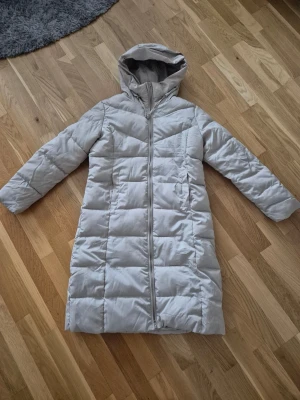 Beige lång dunjacka med huva - Lång, beige dunjacka med huva och dragkedja framtill. Jackan har quiltad design och raka ärmar, perfekt för kalla dagar. Enkel och stilren look som passar till det mesta. Helt ny endast provad, säljs pga viktuppgång. 