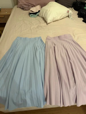 Plisserade kjolar i pastellblå och lila - Två snygga plisserade långkjolar i pastellblått och ljuslila. Kjolarna har bred linning och ett elegant fall med tydliga veck. Perfekta för dig som vill ha en stilren och trendig look.