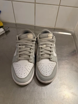 Nike Dunk Low grå och vita sneakers - Nike Dunk Low sneakers i stilren grå och vit färgkombination. Skorna har klassisk låg siluett, perforerad tåbox och ikonisk Swoosh på sidorna. Ovandelen är i läder med matchande grå skosnören och vit mellansula. Perfekta för en clean och modern streetstil.