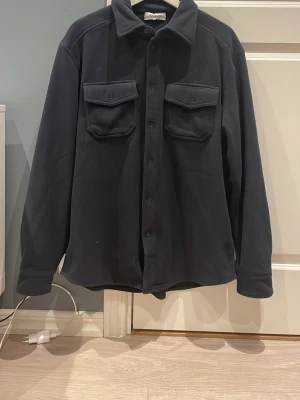 Sandö overshirt marinblå - Stilren svart overshirt från Sandö med klassisk krage, knäppning fram och två bröstfickor. Overshirten har långa ärmar och är tillverkad i ett mjukt, lite tjockare material som ger en avslappnad look. Storlek 50/Medium