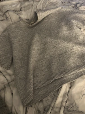 Grå stickad tröja Gina Tricot - Mysig grå stickad tröja från Gina Tricot med kort ärm och rund halsringning. Tröjan har en rak passform och ribbstickade detaljer vid ärmslut, hals och nederkant. Perfekt att styla lager på lager under kyliga dagar.