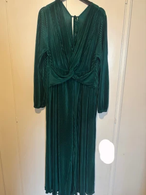 Grön plisserad jumpsuit  - Snygg grön jumpsuit med plisserat tyg och lång ärm. Klänningen har omlott-detalj framtill, knytning i nacken och ett elegant fall. 