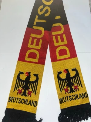 Tysklands halsduk med örn och text - Snygg halsduk i svart, rött och gult med Tysklands örn och texten 'DEUTSCHLAND' i gult. Perfekt accessoar för att visa ditt stöd för Tyskland. Halsduken har fransar i ändarna och är stickad i mjukt material.
