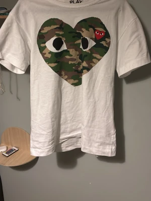 Cdg tisha  - Vit t-shirt från Comme des Garçons Play med stort hjärta i camouflage-mönster och ett litet rött hjärta på bröstet. Klassisk passform och rund halsringning. Perfekt för dig som gillar streetwear och unika prints.