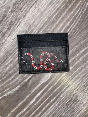 Svart kortfodral med orm från Gucci - Säljer ett svart kortfodral från Gucci med en ikonisk röd, vit och svart orm på framsidan. Fodralet har flera kortfack och är gjort i ett tåligt material med canvasstruktur. Perfekt för dig som vill ha något stilrent men ändå vågat.