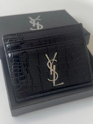 YSL korthållare i krokodilpräglat läder - Snygg svart korthållare från Yves Saint Laurent i glansigt läder med krokodilpräglad yta. Framsidan har det ikoniska YSL-logotypen i silver. Perfekt för kort och enkel att ha i fickan. Helt oanvänd (ny). Kan gå ner i pris vid snabb affär. Skriv vid minsta lilla fundering!