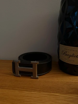 Svart skärp med H-spänne - Snyggt svart bälte med ett stort H-format metallspänne och vita kontrastsömmar. Perfekt accessoar för att lyfta din outfit med en stilren och modern touch. Passar till både jeans och kostymbyxor. 105cm😁