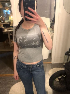 Grå croppad t-shirt med tryck - Snygg grå croppad t-shirt med svart/vitt tryck på bröstet. T-shirten har rund halsringning och korta ärmar. Perfekt för en avslappnad och trendig stil.