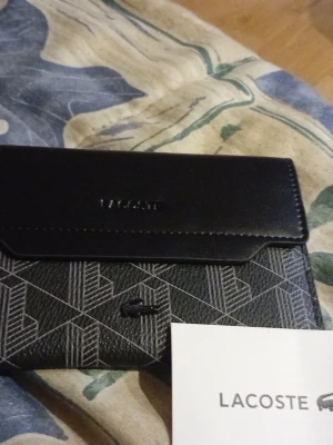 Svart plånbok från Lacoste - Snygg svart plånbok från Lacoste med geometriskt mönster och klassisk krokodil-logga framtill. Plånboken har en stilren design med lock och präglad Lacoste-text. Tillverkad i konstläder med detaljer i svart och grått.