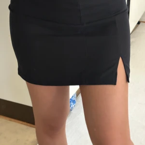 Svart kortkjol med slits - Stilren svart kortkjol med en liten slits framtill på ena sidan. Kjolen har en tight passform och är perfekt för att skapa en trendig look. Tillverkad i ett mjukt och stretchigt material som sitter snyggt på kroppen.