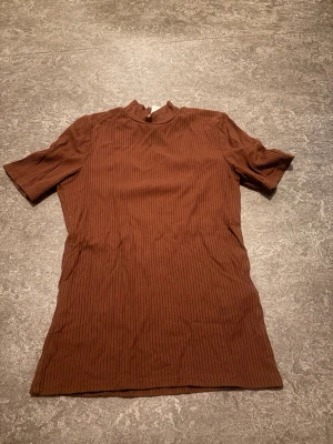 Brun ribbad topp med kort ärm - Snygg brun ribbad topp med korta ärmar och hög hals. Perfekt till jeans eller kjol för en trendig look. Materialet känns mjukt och stretchigt, vilket gör den bekväm att bära hela dagen.