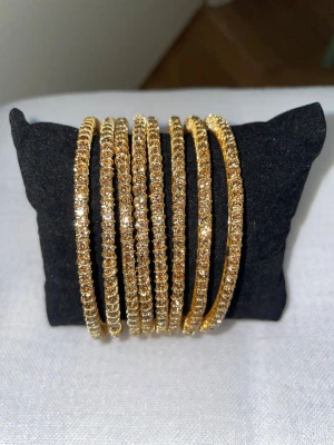Guldiga armband med glittrande stenar - Säljer ett set med åtta tunna armband i guldfärg, dekorerade med gnistrande stenar runt om. Armbanden har en rund form och ger en riktigt lyxig och trendig känsla. Perfekta för att stapla flera tillsammans för en extra statement-look.
