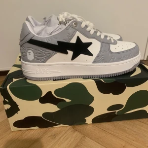 BAPE STA grå/vita sneakers - Säljer ett par BAPE STA sneakers i grått och vitt med svart stjärndetalj på sidan. Skorna har klassisk siluett, perforerad tå, snörning med metall-detaljer och präglade BAPE-loggor. Tillverkade i skinn och levereras med dustbag.