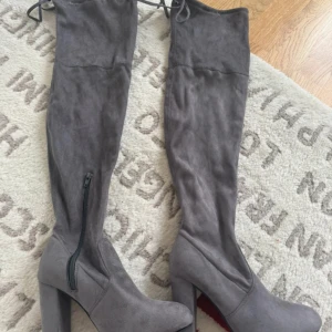 Grå overknee stövlar med klack - Snygga grå overknee stövlar i mockaimitation med hög klack och rund tå. Stövlarna har dragkedja på insidan och snörning upptill för bästa passform. Perfekta för att lyfta vilken outfit som helst.