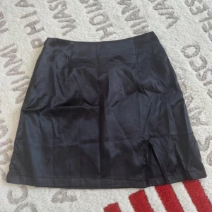 Svart satin kortkjol med slits - Snygg svart kortkjol i satin med en liten slits framtill för extra edge. Kjolen har en stilren och enkel design som gör den lätt att matcha med olika toppar. Perfekt för dig som gillar en clean look med lite attityd.