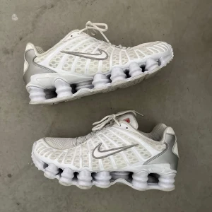 Nike Shox tl vita sneakers med silverdetaljer - Nike Shox sneakers i vitt med silvriga detaljer och ikonisk fjädrande sula. Ovandelen är i mesh och syntet med snörning och silverfärgad Swoosh. Röd Nike-logga på plösen och hälen. Perfekta för dig som vill sticka ut med futuristisk vibe.