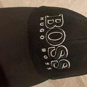 Svart keps från Hugo Boss - Snygg svart keps från Hugo Boss med stor, vit broderad logga framtill. Justerbar rem med metallspänne och vit detalj baktill. Klassisk böjd skärm och ventilerande hål upptill. Perfekt för dig som vill ha en stilren och trendig accessoar.