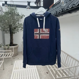 Blå hoodie från Napapijri med norsk flagga - Mörkblå hoodie från Napapijri med broderad norsk flagga och siffran 87 på bröstet. Tröjan har huva med vita snören och en stor magficka framtill. Perfekt för dig som gillar streetwear och snygga detaljer.