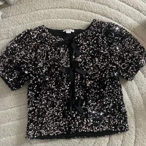Svart paljett bolerotopp Gina Tricot - Glittrig svart bolerotopp från Gina Tricot, täckt med silvriga paljetter och tre svarta rosetter framtill. Kortärmad och croppad modell som ger en festlig vibe. Perfekt till nyår.