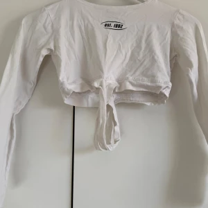Vit croppad långärmad topp med knyt - Säljer en vit croppad långärmad topp med trycket 'est. 1992' i svart framtill. Toppen har knytband i midjan för en snygg detalj och är tillverkad i mjuk bomull. Perfekt för dig som gillar stilrena och enkla plagg med en twist.