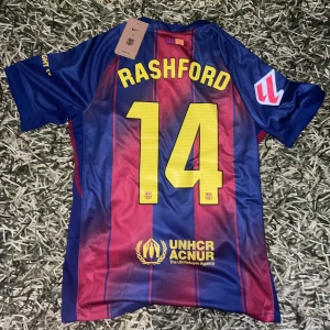 FC Barcelona Rashford #14  - Säljer en FC Barcelona fotbollströja med Rashford och nummer 14 på ryggen. Tröjan har klassiska blå och röda vertikala ränder, gula detaljer och klubbmärke på bröstet. Materialet är lätt och andas, perfekt för match eller träning.