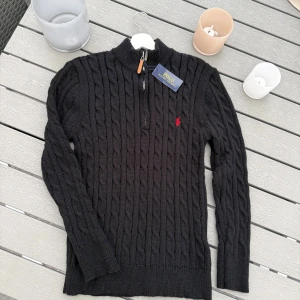 Svart stickad tröja Polo Ralph Lauren - Svart kabelstickad tröja från Polo Ralph Lauren med röd broderad logga på bröstet. Tröjan har half zip med läderdetalj, ribbade muddar och hög krage. Perfekt för dig som gillar klassisk stil med en twist.