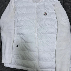 Vit quiltad Moncler pufferjacka - Säljer en stilren vit pufferjacka från Moncler med quiltad framsida och stickade ärmar. Jackan har dragkedja framtill, Moncler-logga på bröstet och en diskret broderad detalj på ärmen. Perfekt för dig som gillar exklusiva märken och cleana vibes.