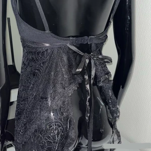 Svart spetstopp med snörning - Supersnygg svart kort klänning i spets med tunna axelband och v-ringning. Klänningen har transparent spetsmaterial med vackert mönster och knytband i ryggen för extra detalj. Perfekt för dig som vill sticka ut med en elegant och edgy look.