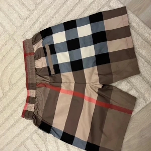 Rutig Burberry-inspirerade shorts - Snygga shorts med klassiskt rutigt mönster i beige, svart, vitt, blått och rött. De har elastisk midja med svart snörning och bakficka. Perfekta för sommaren och ger en clean och trendig look.