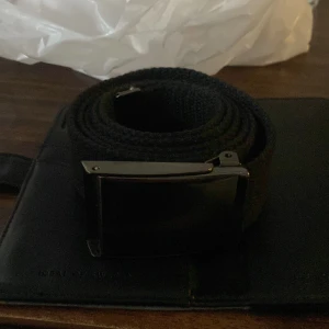 Svart tygbälte med metallspänne - Säljer ett stilrent svart bälte i grovvävt tyg med ett snyggt, rektangulärt metallspänne i silverfärg. Bältet är enkelt att justera och passar perfekt till jeans eller chinos. Minimalistisk design som funkar till många olika outfits.