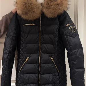 Svart pufferjacka Rockandblue med päls - Svart pufferjacka från Rockandblue med stor fluffig päls i brunt runt kragen. Jackan har guldfärgade dragkedjor, flera fickor och ett Rockandblue-märke på ärmen. Quiltad design på sidorna och ärmarna ger en snygg look. Perfekt för kalla dagar. Fattas en knapp mellan päls och luva, men inget man märker av. Nypris: 3000