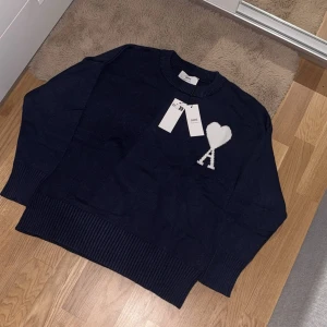 Mörkblå stickad tröja från Ami - Snygg mörkblå stickad tröja från Ami med rund hals och ribbade muddar. På bröstet finns ett vitt hjärta med bokstaven A, som ger en unik och trendig vibe. Perfekt för dig som gillar stilrena detaljer och vill sticka ut lite extra.