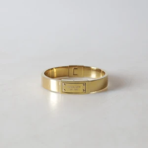 Guldigt armband från Michael Kors - Stilrent armband i guldton från Michael Kors med en rektangulär platta framtill där märkets namn och årtal är ingraverat. Armbandet har en enkel, modern design med diskret lås och passar perfekt för dig som gillar klassiska accessoarer.