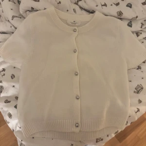 Vit kortärmad kofta från H&M - Superfin vit kofta från H&M med korta ärmar och rund hals. Den har silvriga knappar framtill och ribbade muddar vid ärmslut och nederkant. Mjuk stickad känsla, perfekt för vår och sommar. Enkel och clean stil som funkar till många outfits.