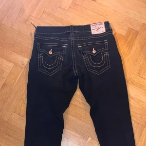 True Religion svarta jeans - Svarta jeans från True Religion med tydliga vita kontrastsömmar och ikoniska fickor bak med lock och knapp. Klassisk femficksmodell, guldiga nitar och knapp, samt patch med logga bak i midjan. Snygg, modern passform som funkar till streetwear. size 36 men passar 34