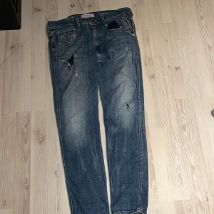 Replay Anbass blå jeans slim fit - Säljer ett par Replay Anbass jeans i blå tvätt med slitningar och hål för en cool, avslappnad look. Slim fit-modell med klassisk femficksdesign och knappgylf. Materialet är jeans och passformen är smal. Perfekt för dig som gillar streetstyle.