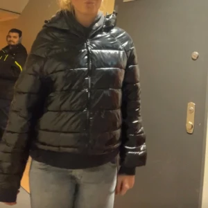 Svart pufferjacka med huva - Säljer en svart, glansig pufferjacka med huva och dragkedja framtill. Jackan har en loose passform och är perfekt för kalla dagar. Den har långa ärmar och ribbad nederkant för extra komfort. Snygg och enkel att matcha med jeans eller andra plagg.